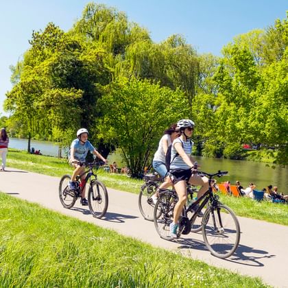 Two cyclists with helmets riding on paved path along Neckar River, surrounded by lush green trees and grass with people relaxing by water.