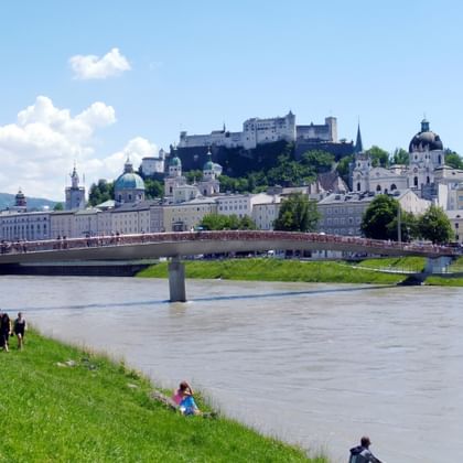 View of Salzburg from the Salzach River showing the historic old town with baroque churches, Hohensalzburg Fortress on the hill, and a bridge.