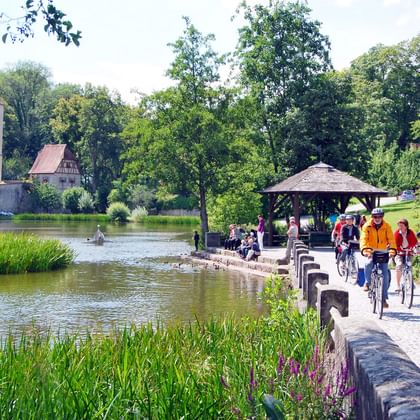 Radfahrer auf Steinbrücke über Teich in Dinkelsbühl mit mittelalterlichem Turm und Pavillon. Grüne Bäume und Schilf umgeben das Wasser.