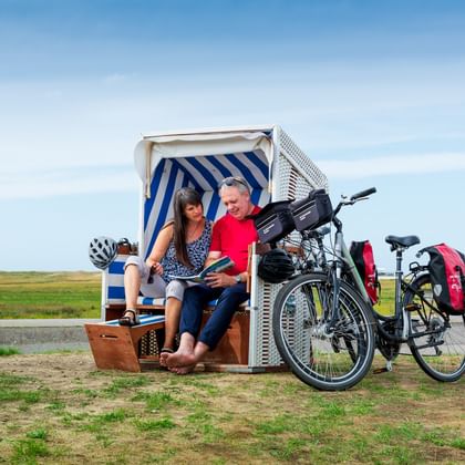 Two cyclists sitting in a blue and white striped beach chair with their touring bikes parked nearby on grassy coastal area at North Sea.