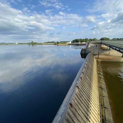 View of the Geesthacht barrage on the Elbe River with metal barrier wall, bridge structure, and calm water reflecting blue sky with clouds.