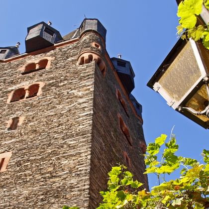 Medieval stone tower in Bernkastel-Kues with red shuttered windows against blue sky. Green grape vines and traditional lantern visible in foreground.