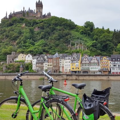 Zwei grüne Fahrräder am Moselufer mit der Reichsburg Cochem auf dem Hügel und bunten Fachwerkhäusern entlang des Flussufers.