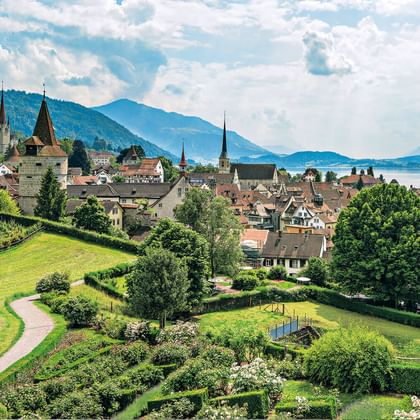 Schweizer Stadt mit Kirchtürmen, rotgedeckten Gebäuden, grünen Gärten und Weinbergen. See und Berge im Hintergrund unter bewölktem Himmel.