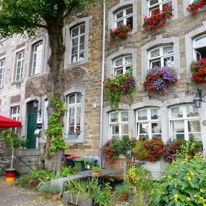 Historic stone buildings in Kornelimünster city centre with colorful flower boxes on windows. Red umbrellas and outdoor seating visible on left.