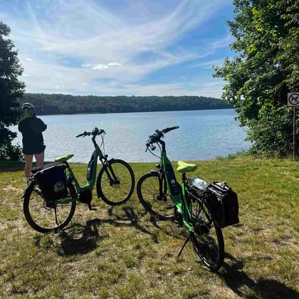 Zwei E-Bikes mit Taschen auf Wiese am Badeplatz Werbellinsee. Eine Person steht am Ufer und blickt auf den See und bewaldete Hügel.
