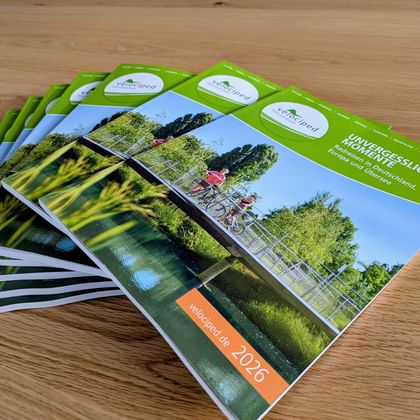 Stack of green Velociped cycling catalogs for 2026 on wooden surface, showing cyclists on tree-lined path with river reflection.