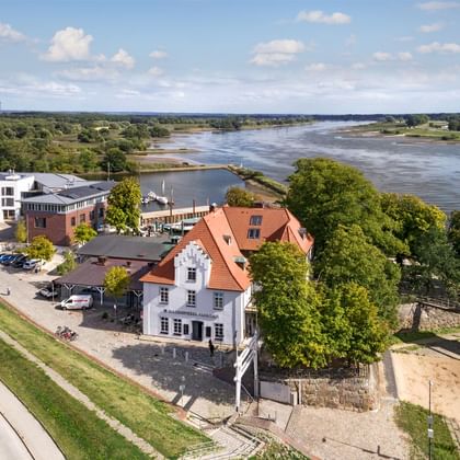 Luftaufnahme des Zollenspieker Fährhauses mit weißem historischem Gebäude und rotem Ziegeldach an der Elbe, umgeben von grünen Bäumen.