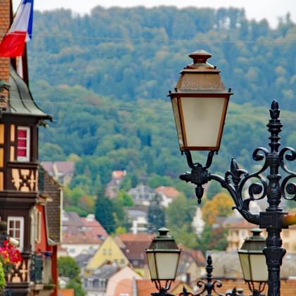 Verzierte schwarze Straßenlaterne mit vier Laternen in Saverne, Frankreich. Historische Fachwerkhäuser und französische Flagge links, bewaldete Hügel im Hintergrund.