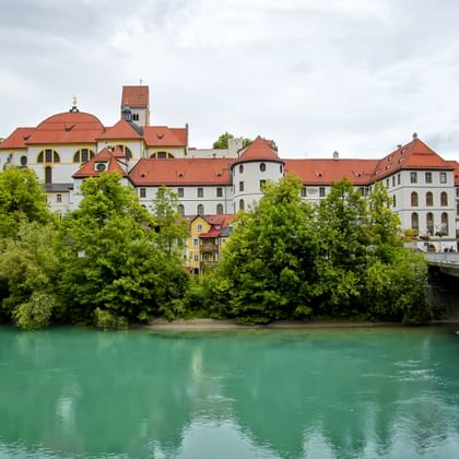 View of Füssen's historic old town with colorful buildings and red-tiled roofs along a turquoise river. A concrete bridge spans the water in the foreground.