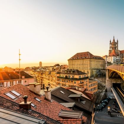 Sunset view over Lausanne rooftops with cathedral towers and a yellow bridge visible. Golden light illuminates the historic buildings.