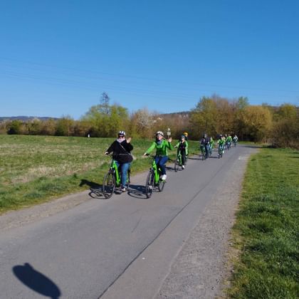 Group of cyclists riding on paved path through green meadows under blue sky. Trees line the path with hills visible in the distance.