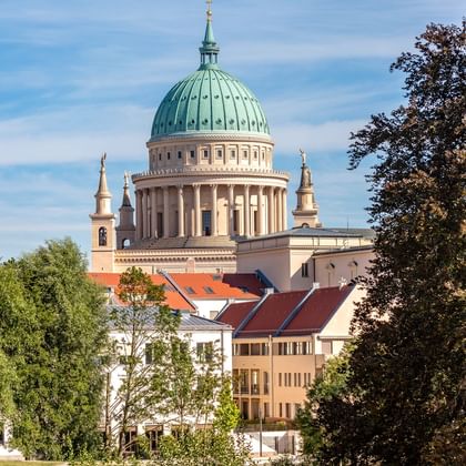 Potsdam's Nikolaikirche with distinctive green copper dome and classical columns, surrounded by trees and residential buildings under blue sky.