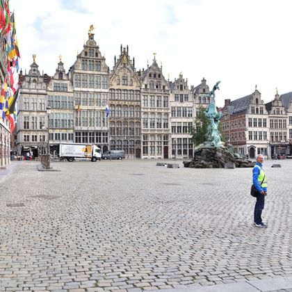 Kopfsteinpflasterplatz in Antwerpen mit verzierten flämischen Gildehäusern, Bronzebrunnen und Person in Warnweste. Bunte Flaggen hängen am Gebäude.