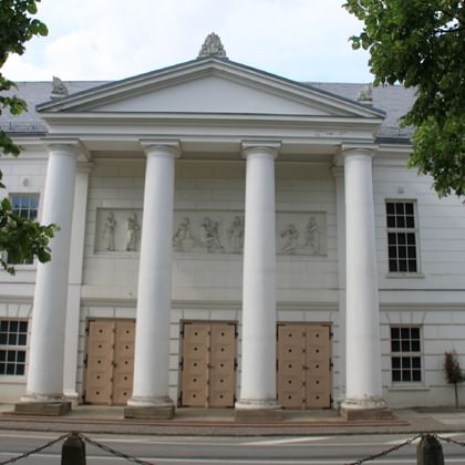 Weißes klassizistisches Gebäude in Putbus mit sechs hohen Säulen, die einen dreieckigen Giebel stützen. Drei Holztüren mit Metallbeschlägen am Eingang.