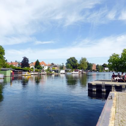 Waterfront promenade in Malchow with calm lake, wooden pier, colorful houses, green trees and blue cloudy sky.