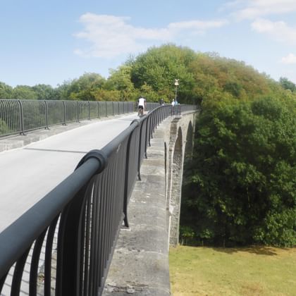 Betonbrücke mit Metallgeländer über den Rurstausee. Zwei Radfahrer sind auf dem Brückenweg sichtbar, umgeben von grünen Bäumen.