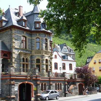 Stone villa with slate roof and tower in Bernkastel-Kues, surrounded by traditional German buildings and vineyard-covered hills.