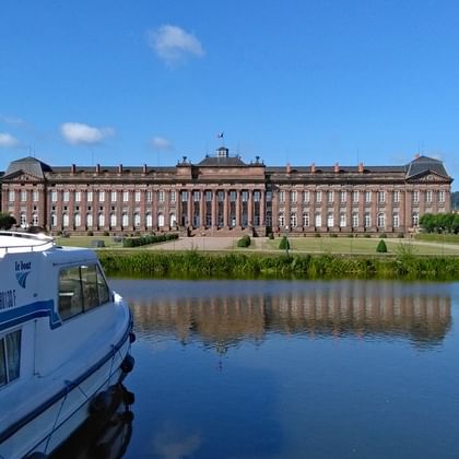 Château des Rohan in Saverne, a large red sandstone palace reflected in the canal. A blue and white boat is moored in the foreground.