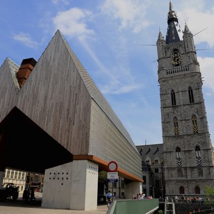 Medieval Belfort bell tower in Ghent next to a modern angular building with wooden facade under blue sky with white clouds.