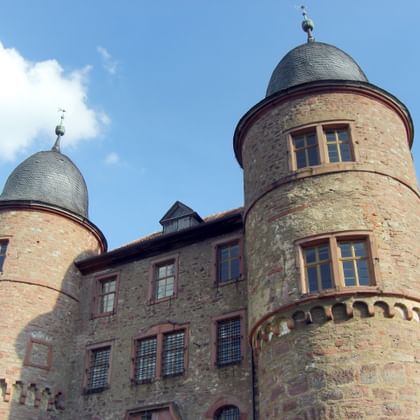 Historische Burg Wertheim mit zwei runden Steintürmen mit dunklen Schieferdächern und einem zentralen Gebäude mit rot gerahmten Fenstern.