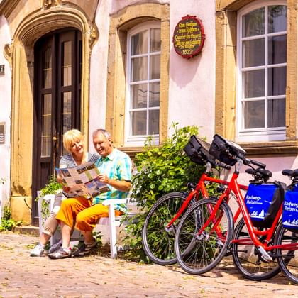Zwei Radfahrer mit Karte ruhen auf weißen Stühlen vor einem historischen Gebäude in Bad Wimpfen. Zwei rote Tourenräder mit blauen Taschen stehen daneben.