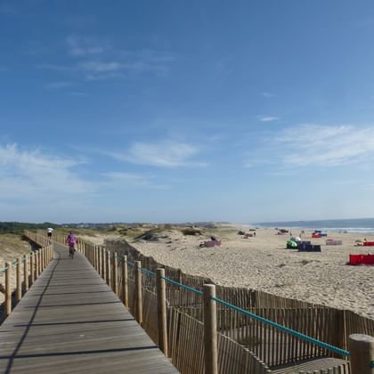 Holzsteg mit Geländer führt zu einem Sandstrand an Portugals Nordküste. Bunte Strandhütten und Sonnenschirme säumen das Ufer unter blauem Himmel.