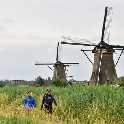 Two cyclists in tall grass with historic Dutch windmills at Kinderdijk in the background under cloudy sky.