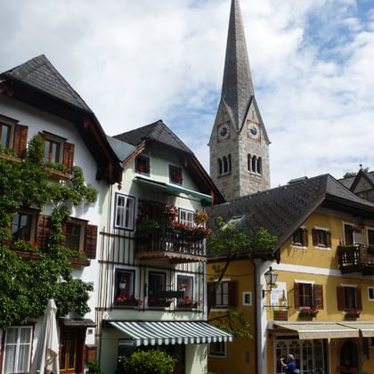 Traditional Austrian houses in Hallstatt with colorful facades and wooden shutters. A tall church spire with clock dominates the skyline.