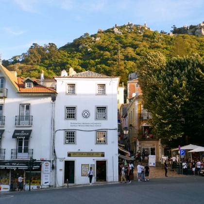 Town square in Sintra with white Portuguese buildings, tourists walking, and forested hills with castle ruins visible in the background.