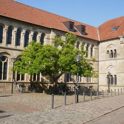 Historic stone buildings with red tile roofs in Osnabrück featuring Romanesque arched windows and columns. A statue stands in the cobblestone square.