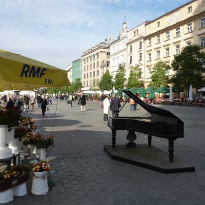 Pedestrian square in Krakau Old Town with black grand piano, flower market stalls under yellow RMF FM umbrella, and historic buildings.
