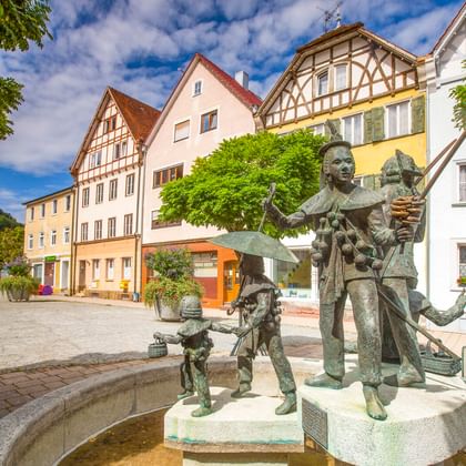 Brunnen mit bronzenen Narrenfiguren auf dem Platz in Oberndorf, umgeben von traditionellen Fachwerkhäusern und grünen Bäumen unter blauem Himmel.