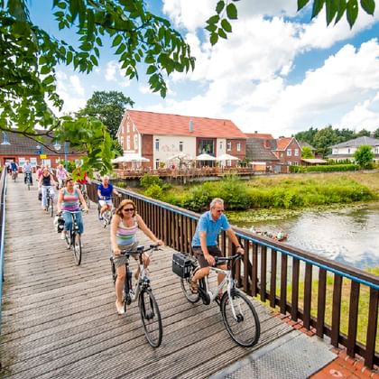Group of cyclists crossing a wooden bridge over a river near Herzlake. Traditional German houses with red roofs visible in background.