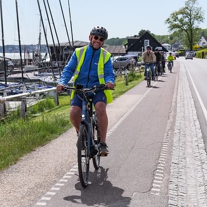 Gruppe von Radfahrern auf einer asphaltierten Straße entlang der Küste. Historisches Segelschiff links, Wasser und Häuser im Hintergrund.