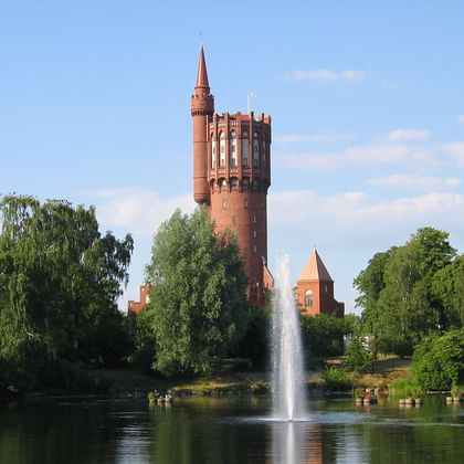 Roter Backsteinwasserturm in Landskrona ragt über grüne Bäume, mit einem Springbrunnen in einem Teich im Vordergrund unter blauem Himmel.