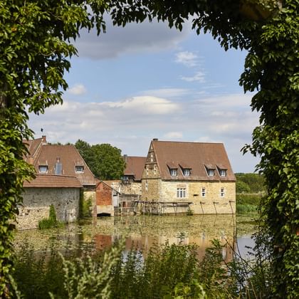 Blick auf Burg Kakesbeck in Lüdinghausen durch grüne Äste, zeigt historische Steingebäude mit roten Ziegeldächern am Wasser.
