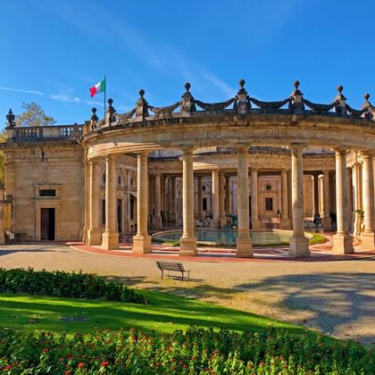 Terme Tettuccio neoclassical spa building with curved colonnade, ornate balustrade, and Italian flag. Manicured gardens with colorful flowers in foreground.