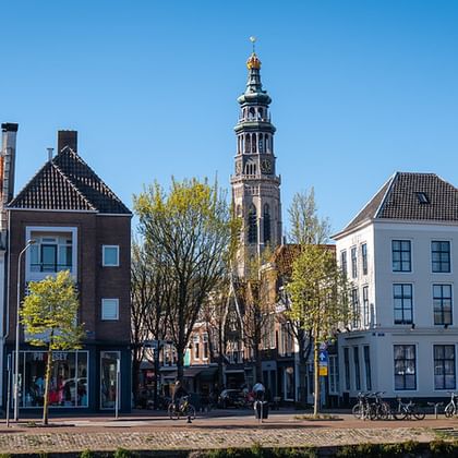 Row of traditional Dutch canal houses with varied gabled facades along a waterfront in Zeeland, with a tall church tower rising behind trees.