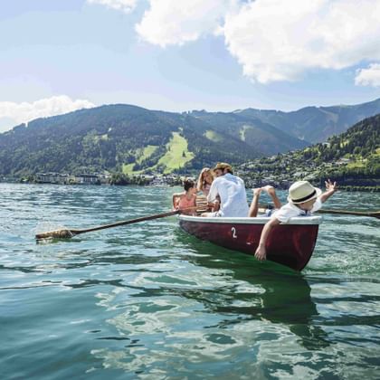 Red rowing boat with five people on turquoise Zeller See. Mountains and green slopes visible in background under partly cloudy sky.