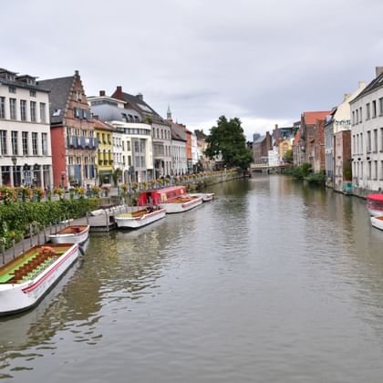 Canal in Ghent lined with colorful historic buildings and tour boats. Traditional Flemish architecture with white, yellow, and red facades.