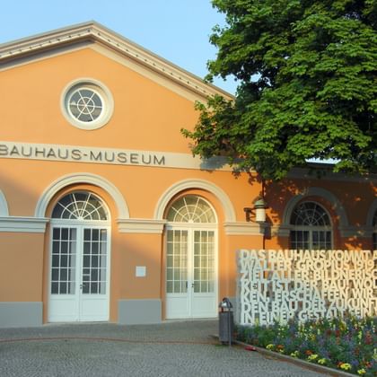 Bauhaus-Museum Weimar orangene Fassade Oranges Bauhaus-Museum Weimar mit weißen Verzierungen, Bogenfenstern, rundem Fenster und Textwand neben buntem Blumengarten.