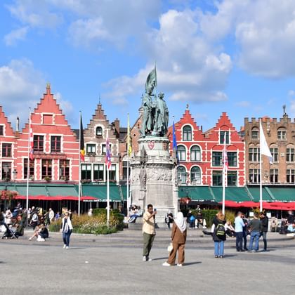 Historischer Grote Markt Platz in Brügge mit bunten mittelalterlichen Gebäuden, zentralem Denkmal und Touristen auf dem Platz.