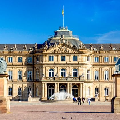 Neues Schloss Stuttgart, ein barockes Schloss mit gelber Fassade, flankiert von zwei Säulen mit Löwenstatuen unter blauem Himmel.