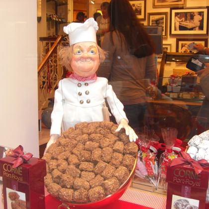 Kochfigur mit Schokoladentrüffeln in Brüssel Dekorative Kochfigur in weißer Uniform und Mütze präsentiert Schokoladentrüffel in einem belgischen Schokoladenladen mit Kunden im Hintergrund.
