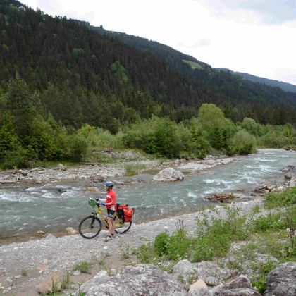 Radfahrer in roter Jacke mit Tourenrad am steinigen Ufer der türkisfarbenen Drau, bewaldete Berge im Hintergrund unter bewölktem Himmel.