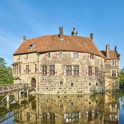 Mittelalterliche Burg Vischering in Lüdinghausen umgeben von Wassergraben, mit Steinmauern, rotem Ziegeldach und Holzbrücke unter blauem Himmel.