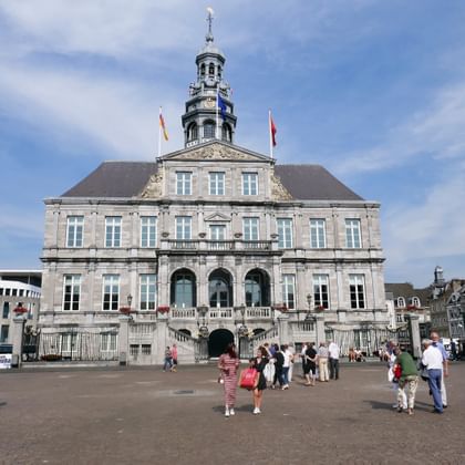 Rathaus Maastricht, ein barockes Steingebäude mit verziertem Turm und Uhr, wehende Flaggen. Touristen versammeln sich auf dem Platz davor.
