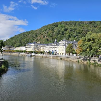 View of Bad Ems with Häcker's Grand Hotel along the Lahn River. Historic buildings line the waterfront with forested hills in the background.