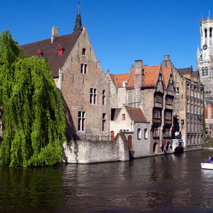 Historic Bruges canal with medieval buildings Medieval buildings line a canal in Bruges with the famous Belfry tower visible. A weeping willow hangs over the water and a small boat carries tourists.